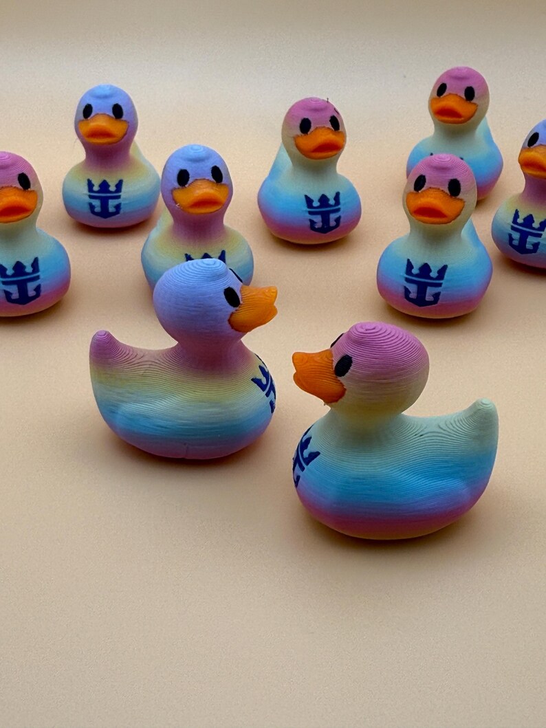 May include: A collection of small, 3D-printed rubber ducks featuring a gradient rainbow color scheme. Each duck has an orange beak, black eyes, and a dark blue anchor-like logo printed on its chest. The ducks are arranged in a group.