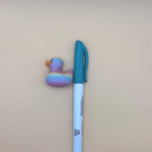 May include: A small rubber duck with a pastel rainbow gradient finish and an orange beak, positioned next to a white Sharpie Creative Marker with a teal cap and base, providing a visual scale reference for the duck's size.