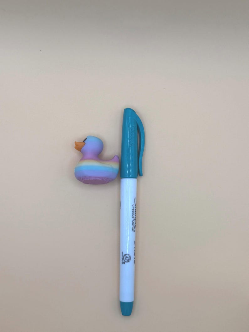 May include: A small rubber duck with a pastel rainbow gradient finish and an orange beak, positioned next to a white Sharpie Creative Marker with a teal cap and base, providing a visual scale reference for the duck's size.