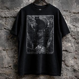 Black gothic t-shirt featuring a dark woodcut-style black cat illustration perched above skulls in a haunted forest with the phrase “Creature of the Night.”