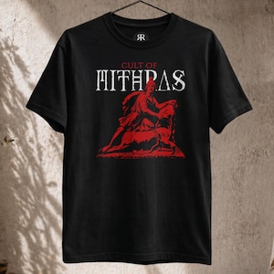May include: A black short-sleeve t-shirt featuring a red graphic print of a figure kneeling over a bull. Above the graphic, the text reads CULT OF MITHRAS in white capital letters. The shirt has a crew neckline and is displayed on a black hanger.