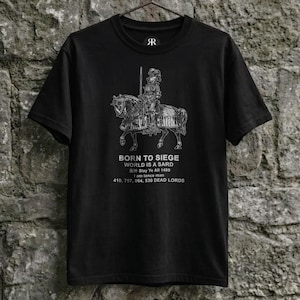 May include: A black short-sleeved t-shirt featuring a white graphic of a knight on horseback. The text reads: BORN TO SIEGE, WORLD IS A SARD, Slay Ye All 1489, I am lance man, 410, 757, 664, 530 DEAD LORDS. The shirt is displayed on a black hanger.
