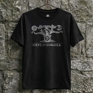 Könnte beinhalten: Schwarzes T-Shirt mit einer detaillierten silbernen Grafik, die Sonne, Mond und drachenartige Figuren zeigt. Der Text "SOLVE ET COAGULA" ist darunter gedruckt. Das T-Shirt hängt an einem Metallbügel vor einer Steinmauer.
