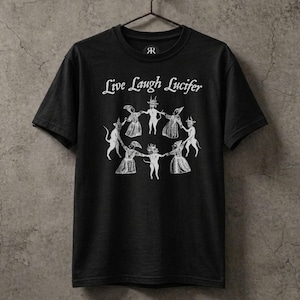 Puede incluir: Camiseta negra con la frase "Live Laugh Lucifer" en blanco, con una fuente gótica. Debajo del texto, una ilustración circular de figuras con capas y máscaras con cuernos. La camiseta está colgada en una percha de metal.