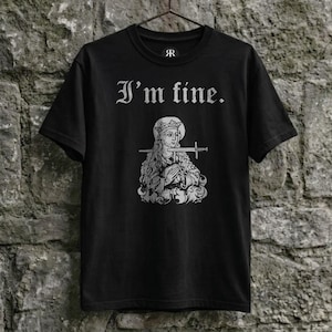 May include: Black t-shirt with the phrase "I'm fine" in gothic font above a detailed illustration of a figure holding a sword. The image is printed in white. The shirt hangs on a metal hanger against a stone wall.