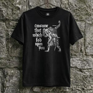 Op de afbeelding: Zwart t-shirt met een gotische illustratie in wit van twee figuren, waarvan één een zwaard hanteert. De tekst "Consume that which fed upon you" is in het wit gedrukt. Het t-shirt hangt aan een metalen hanger.