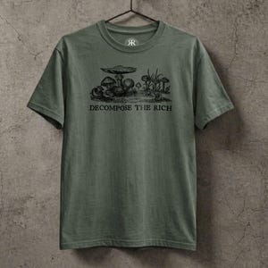 Olive green graphic t-shirt featuring a vintage mushroom illustration with the phrase “Decompose the Rich” in black lettering, displayed on a hanger against a textured stone wall in a dark earthy aesthetic.