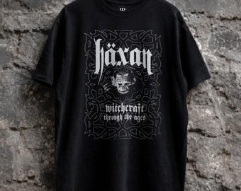 May include: Black t-shirt with the word "häxan" in a gothic font, a skull graphic, and the text "witchcraft through the ages." The design is in white. The shirt is hanging on a black hanger against a textured gray brick wall.