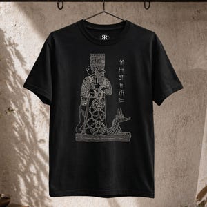 Peut inclure: T-shirt noir avec un motif blanc représentant une figure avec une haute coiffe, un bâton et une créature ressemblant à un chien. Le t-shirt porte les lettres "R" sur le haut du côté gauche de la poitrine. Le motif comprend du texte vertical sur le côté droit de la figure.