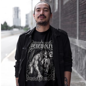 May include: A black t-shirt featuring a white graphic print of a werewolf holding a lamb under a full moon, with the text "WEREWOLF" arched above the illustration. The shirt is worn under a black button-up jacket with a structured collar and pockets.