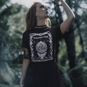 May include: A woman wearing a black t-shirt with a white graphic design that says "Theosophia" and a black skirt. She is standing in a forest and has her hand raised in the air.