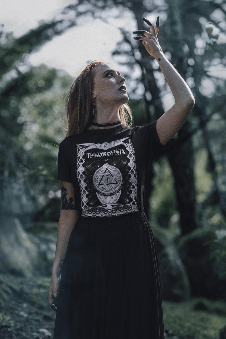 May include: A woman wearing a black t-shirt with a white graphic design that says "Theosophia" and a black skirt. She is standing in a forest and has her hand raised in the air.
