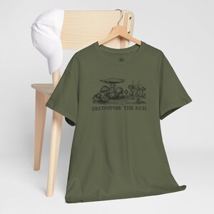May include: Olive green t-shirt with a black illustration of mushrooms and the text "DECOMPOSE THE RICH." The shirt is hanging on a wooden hanger, with a white baseball cap resting on a wooden chair.