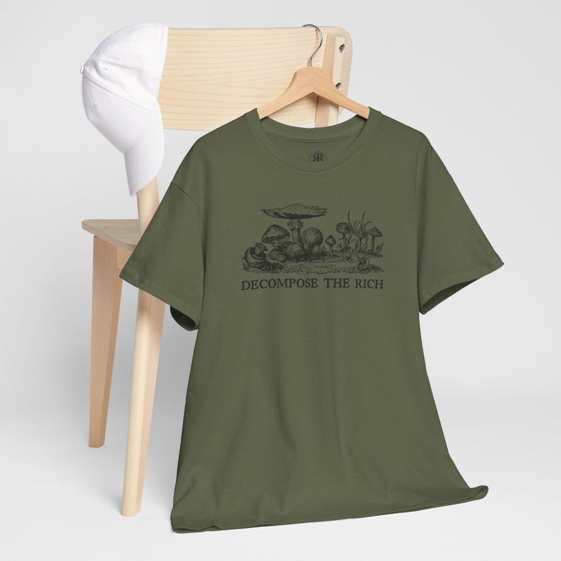 May include: Olive green t-shirt with a black illustration of mushrooms and the text "DECOMPOSE THE RICH." The shirt is hanging on a wooden hanger, with a white baseball cap resting on a wooden chair.