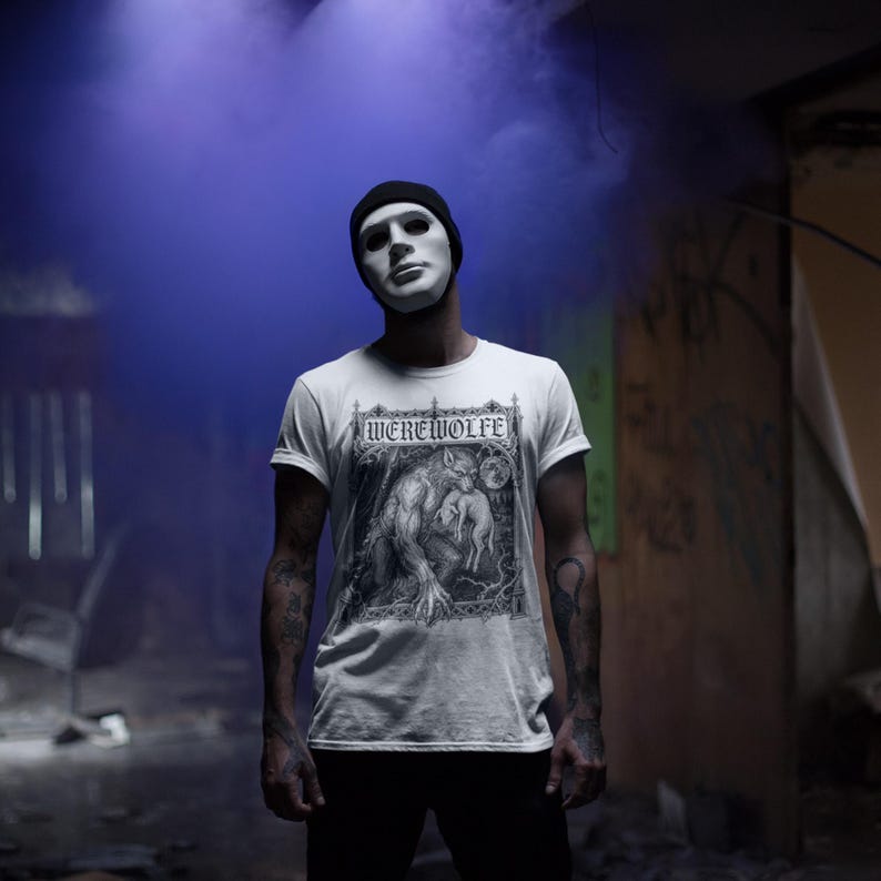 May include: A white short-sleeved t-shirt featuring a black and white graphic print of a werewolf holding a small animal under the text WEREWOLFE. The shirt is worn with a black beanie and black pants against a backdrop of purple smoke.