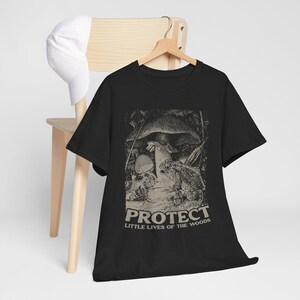 May include: Black t-shirt with a nature-themed graphic. The design features a mushroom, frog, and other forest elements, with the text "PROTECT LITTLE LIVES OF THE WOODS" in a bold font. A white baseball cap hangs on a wooden chair.