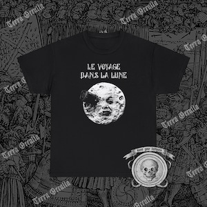 以下が含まれることがあります： 月の男の顔の白いグラフィックが描かれた黒いTシャツ。男は望遠鏡をのぞいています。グラフィックの上に「Le Voyage Dans La Lune」というテキストがあります。グラフィックの下に「Know your darkness」というテキストがあります。