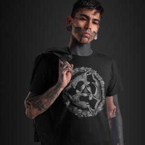 May include: A black short-sleeved t-shirt featuring a circular graphic of three hares arranged in a triangle, surrounded by a detailed wreath of oak leaves and acorns, with a small triquetra symbol positioned in the center of the hares.