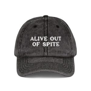 May include: A distressed black baseball cap with a curved brim. The cap features the white text "ALIVE OUT OF SPITE" across the front. The hat has a vintage, worn look.