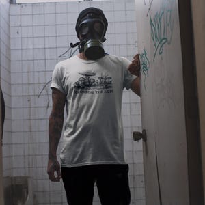 May include: A light gray t-shirt with a black graphic of mushrooms and the text "DECOMPOSE THE RICH". The person is wearing a black gas mask and a black beanie. The background is a tiled wall and a door with graffiti.