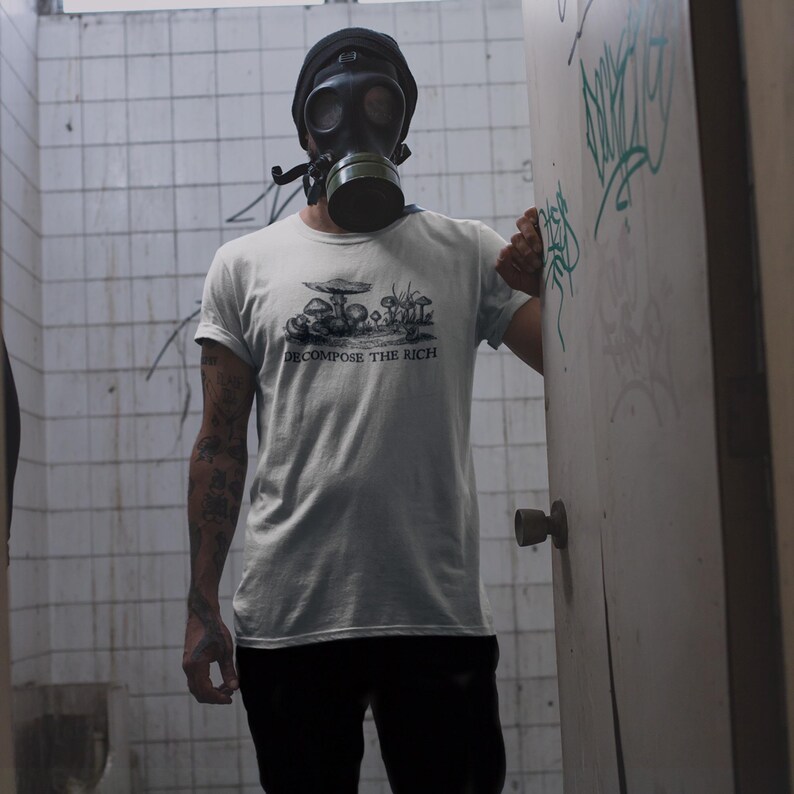 May include: A light gray t-shirt with a black graphic of mushrooms and the text "DECOMPOSE THE RICH". The person is wearing a black gas mask and a black beanie. The background is a tiled wall and a door with graffiti.