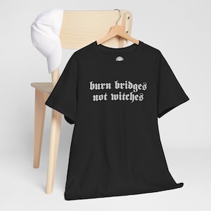 May include: A black short-sleeved t-shirt featuring the white text "burn bridges not witches" in a gothic-style font printed across the chest. The garment is displayed on a wooden hanger, with a plain white baseball cap resting nearby.