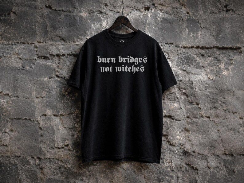 Black Burn Bridges Not Witches shirt with gothic blackletter text printed across the chest, shown hanging against a stone wall.