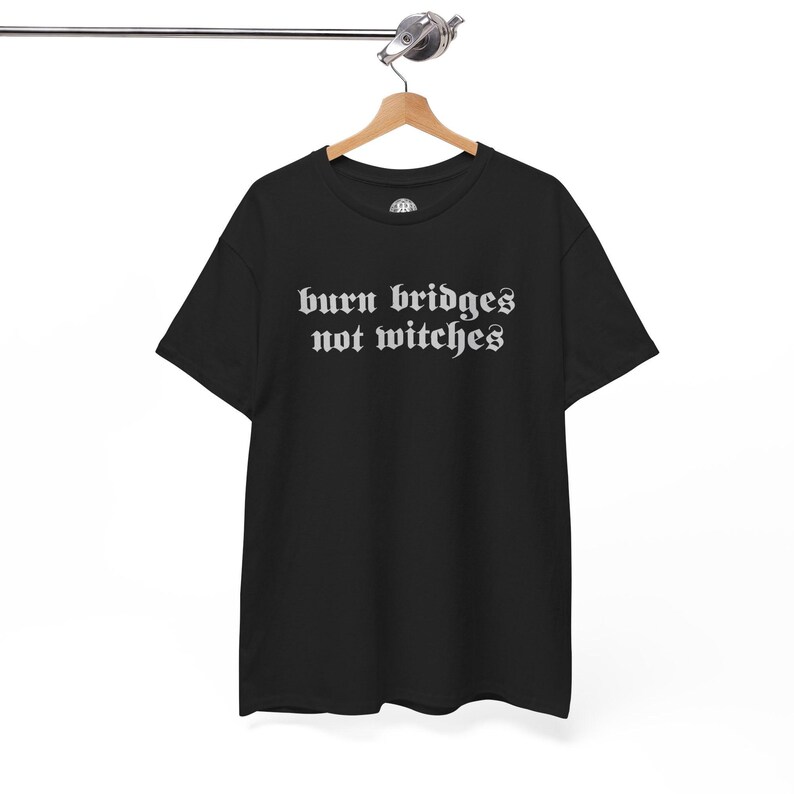May include: A black short-sleeve t-shirt featuring the white text "burn bridges not witches" printed in a gothic-style font across the chest. The garment has a crew neckline and is displayed on a wooden hanger against a plain white background.