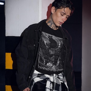 Pu&ograve; includere: T-shirt nero con una grafica dettagliata in bianco e nero di un gatto in una foresta, con la scritta "CREATURE OF THE NIGHT". La maglietta &egrave; indossata con una giacca di jeans nera e una camicia a quadri in bianco e nero.