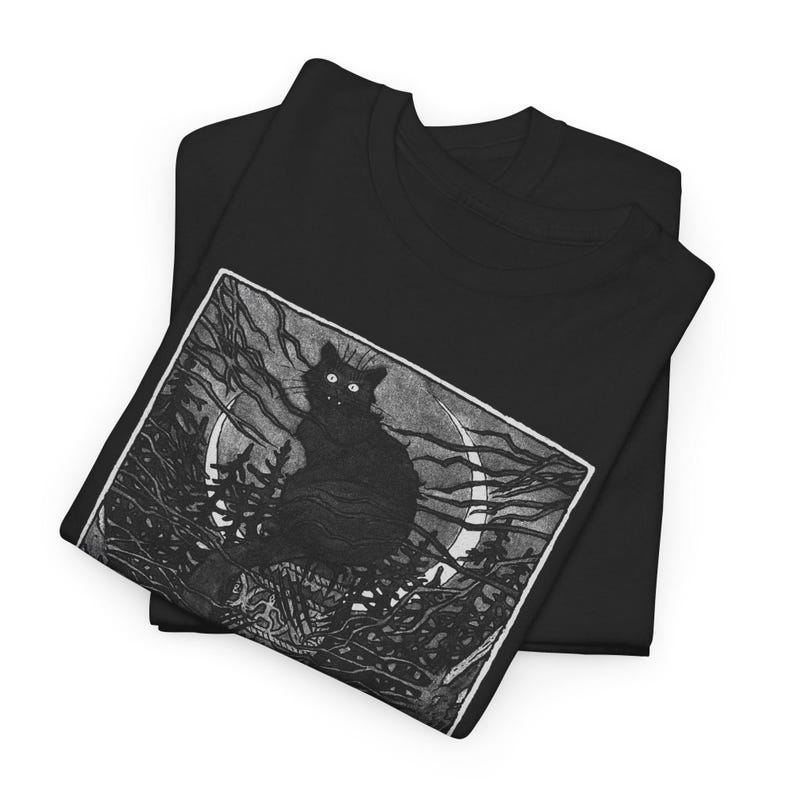 Pu&ograve; includere: T-shirt nero con una grafica di un gatto nero con occhi luminosi, appollaiato su un albero contro una luna crescente. Il design &egrave; in stile xilografia vintage. La maglietta &egrave; piegata, mostrando il disegno sopra.