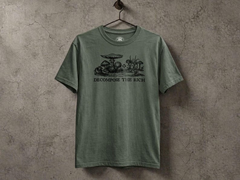Olive green graphic t-shirt featuring a vintage mushroom illustration with the phrase “Decompose the Rich” in black lettering, displayed on a hanger against a textured stone wall in a dark earthy aesthetic.