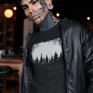 Puede incluir: Una camiseta negra con un gr&aacute;fico blanco de una silueta de bosque. El gr&aacute;fico tiene un aspecto desgastado y vintage. La camiseta la lleva puesta una persona con tatuajes de tinta negra.