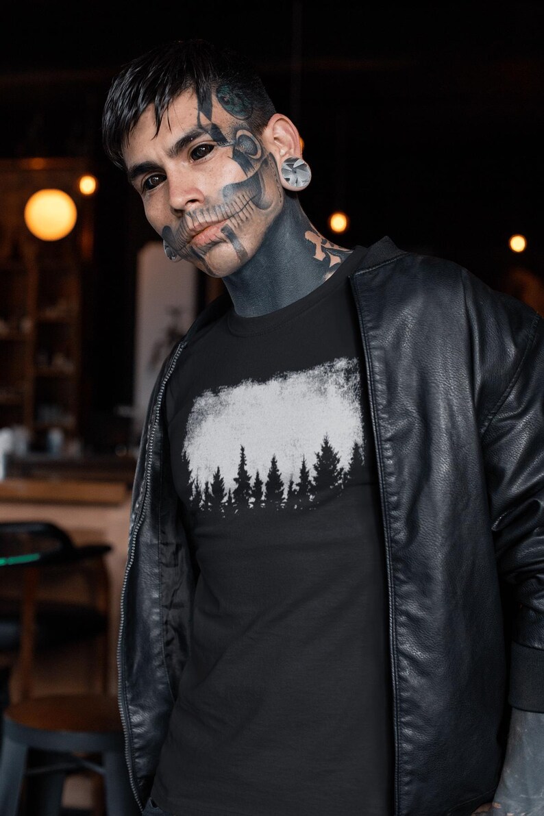 Puede incluir: Una camiseta negra con un gr&aacute;fico blanco de una silueta de bosque. El gr&aacute;fico tiene un aspecto desgastado y vintage. La camiseta la lleva puesta una persona con tatuajes de tinta negra.