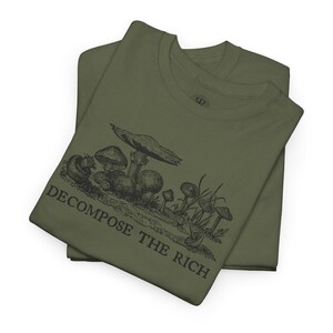May include: Two olive green t-shirts are stacked, displaying a black illustration of various mushrooms and the text "DECOMPOSE THE RICH." The graphic features detailed drawings of different mushroom types, with the text below in a bold, sans-serif font.