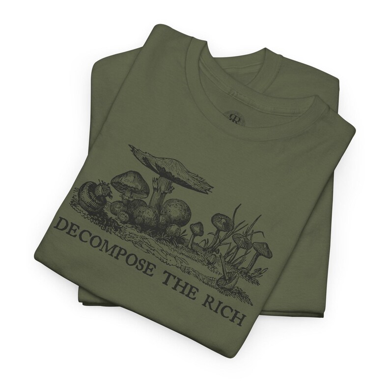 May include: Two olive green t-shirts are stacked, displaying a black illustration of various mushrooms and the text "DECOMPOSE THE RICH." The graphic features detailed drawings of different mushroom types, with the text below in a bold, sans-serif font.