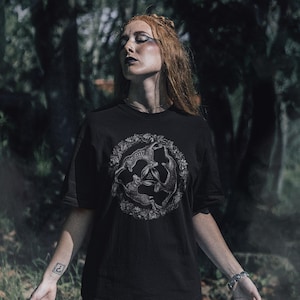 May include: A black short-sleeved t-shirt featuring a circular graphic print of three hares arranged in a triangular formation, surrounded by a detailed wreath of leaves and flowers, with a small triquetra symbol centered between the hares.