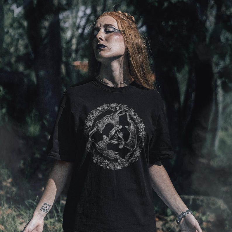 May include: A black short-sleeved t-shirt featuring a circular graphic print of three hares arranged in a triangular formation, surrounded by a detailed wreath of leaves and flowers, with a small triquetra symbol centered between the hares.