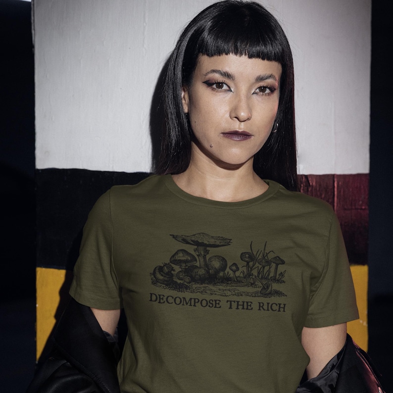 May include: Olive green t-shirt featuring a black illustration of mushrooms and the text "DECOMPOSE THE RICH". The shirt is made of a soft material and has a crew neck.