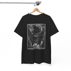 Pu&ograve; includere: T-shirt nero con un'illustrazione in bianco e nero di un gatto nero in una foresta sotto la luna piena. Il design include il testo "CREATURE OF THE NIGHT". La maglietta &egrave; appesa a una gruccia di legno.