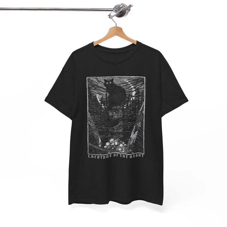 Pu&ograve; includere: T-shirt nero con un'illustrazione in bianco e nero di un gatto nero in una foresta sotto la luna piena. Il design include il testo "CREATURE OF THE NIGHT". La maglietta &egrave; appesa a una gruccia di legno.