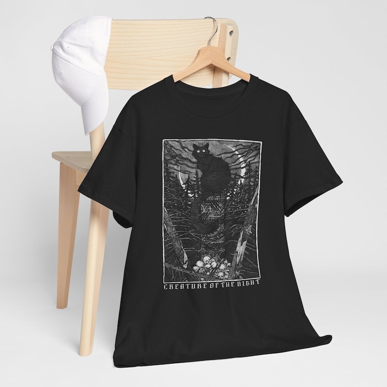 Pu&ograve; includere: T-shirt nero con un'illustrazione in bianco e nero di un gatto nero in una foresta sotto la luna piena. Il design include la scritta "CREATURE OF THE NIGHT". Un berretto da baseball bianco &egrave; appoggiato su una sedia di legno.