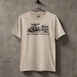 Beige graphic t-shirt featuring a vintage mushroom illustration with the phrase “Decompose the Rich” in black lettering, displayed on a hanger against a textured stone wall in a dark earthy aesthetic.