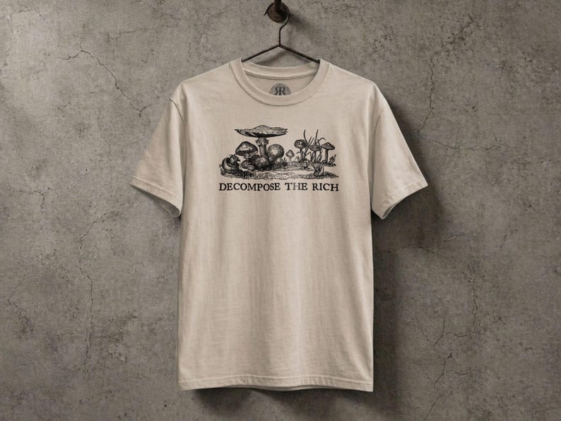 Beige graphic t-shirt featuring a vintage mushroom illustration with the phrase “Decompose the Rich” in black lettering, displayed on a hanger against a textured stone wall in a dark earthy aesthetic.
