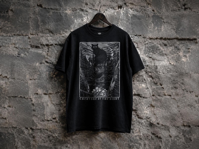 Black gothic t-shirt featuring a dark woodcut-style black cat illustration perched above skulls in a haunted forest with the phrase “Creature of the Night.”