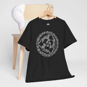 May include: A black short-sleeve t-shirt featuring a white circular graphic of three hares arranged in a triangular formation, surrounded by a detailed leaf border and a small central triquetra symbol, displayed on a wooden hanger.