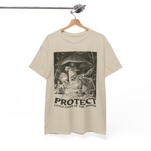 May include: A beige t-shirt featuring a black and white illustration of a frog, mushroom, and skull. The text "PROTECT LITTLE LIVES OF THE WOODS" is printed below the illustration. The shirt is hanging on a wooden hanger.