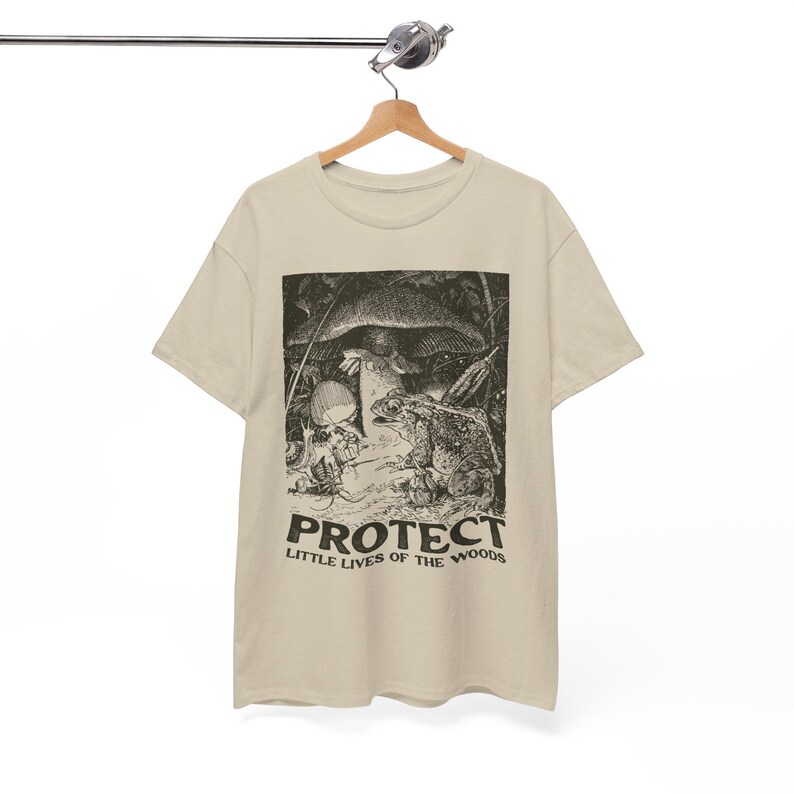 May include: A beige t-shirt featuring a black and white illustration of a frog, mushroom, and skull. The text "PROTECT LITTLE LIVES OF THE WOODS" is printed below the illustration. The shirt is hanging on a wooden hanger.