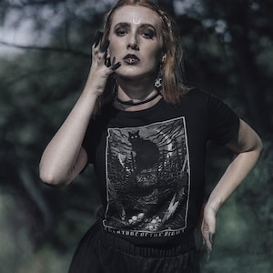 Pu&ograve; includere: T-shirt nera con una grafica di un gatto nero in una foresta e la scritta "CREATURE OF THE NIGHT". La maglietta a maniche corte ha uno scollo tondo. L'immagine &egrave; in bianco e nero.