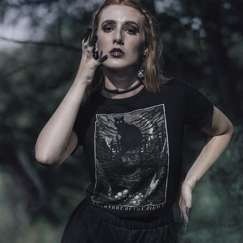 Pu&ograve; includere: T-shirt nera con una grafica di un gatto nero in una foresta e la scritta "CREATURE OF THE NIGHT". La maglietta a maniche corte ha uno scollo tondo. L'immagine &egrave; in bianco e nero.