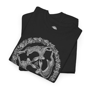May include: A black t-shirt featuring a white graphic print of three hares arranged in a circular formation. The hares are surrounded by a detailed wreath of leaves and acorns, with a small triquetra symbol positioned in the center.